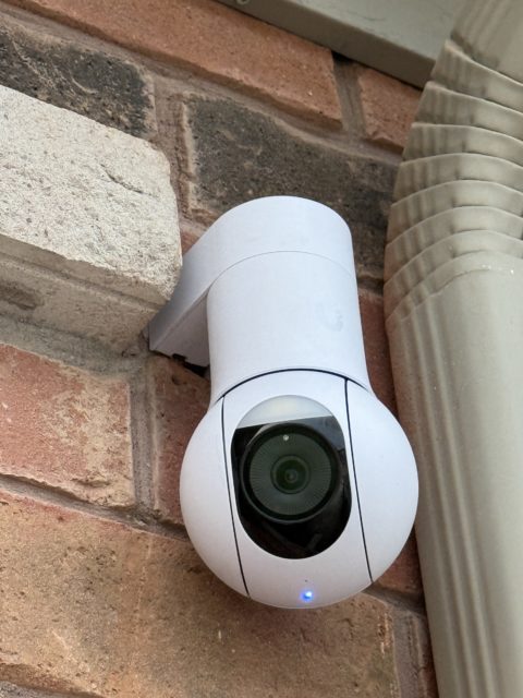 White UniFi security camera mounted on exterior brick beside a downspout
